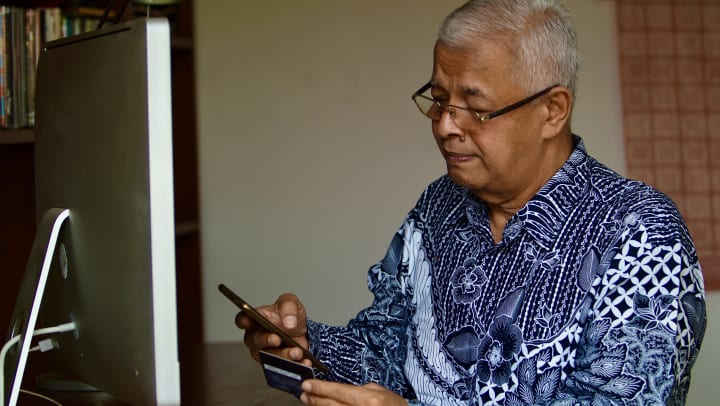 elderly man holding credit card and phone in front of computer